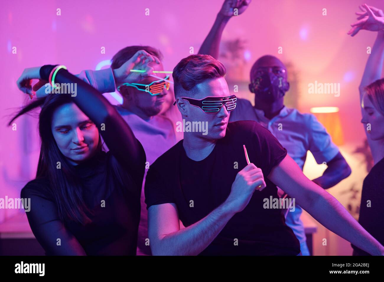 Jeune homme excité en vêtements décontractés et Disco lunettes et son amis interculturels avec bâtons lumineux dansant ensemble tout en appréciant la maison fête Banque D'Images