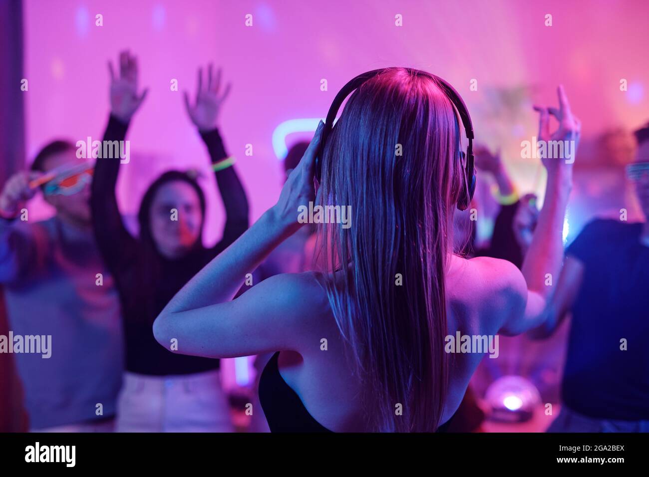 Vue arrière d'une jeune femme avec de longs cheveux blonds en contact un casque tout en se tenant devant la foule d'amis enthousiastes danse à la fête Banque D'Images
