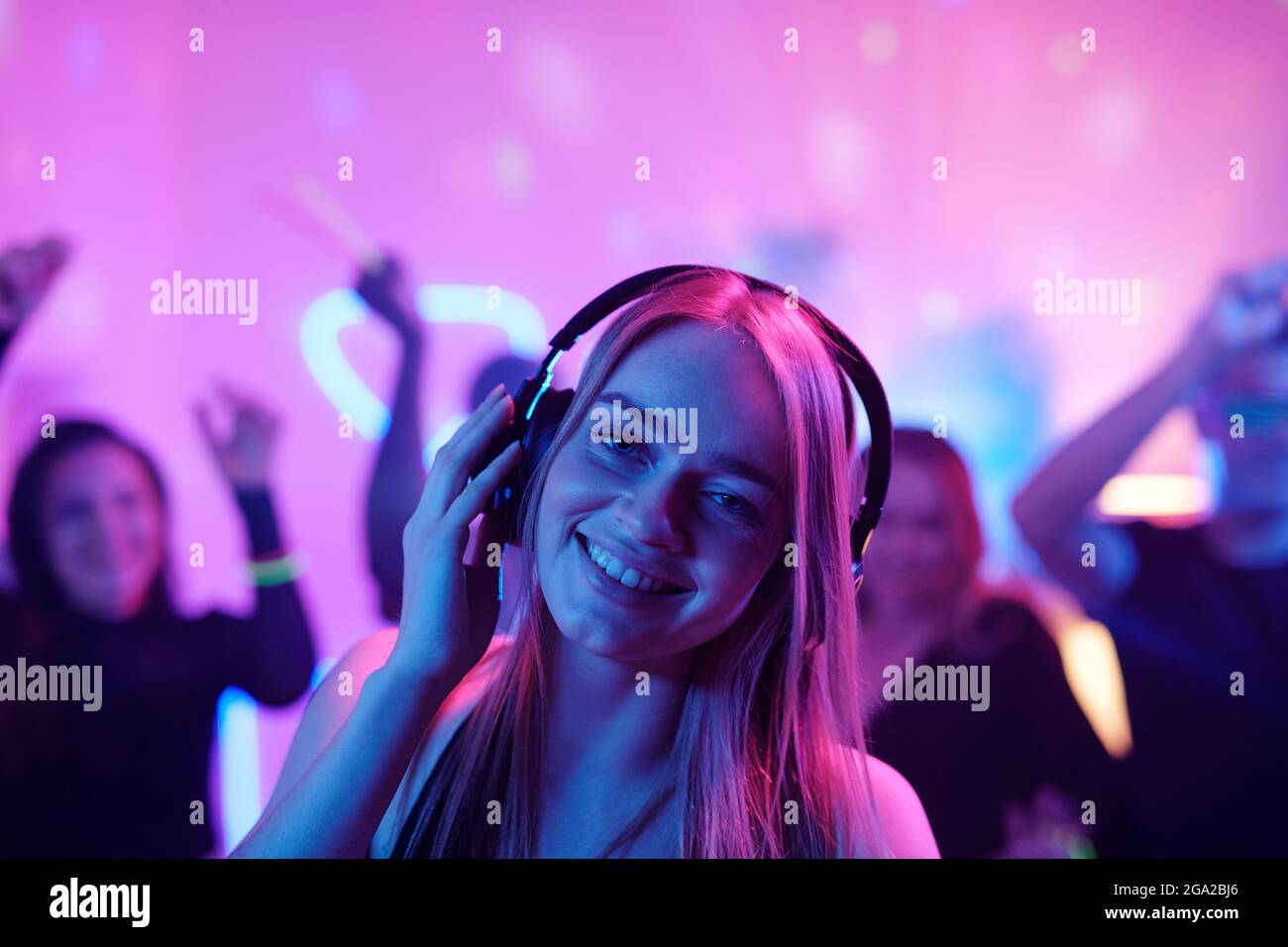 Jeune femme gaie avec de longs cheveux blonds se prenant les doigts pendant écouter de la musique dans un casque devant la foule dansante à fête Banque D'Images