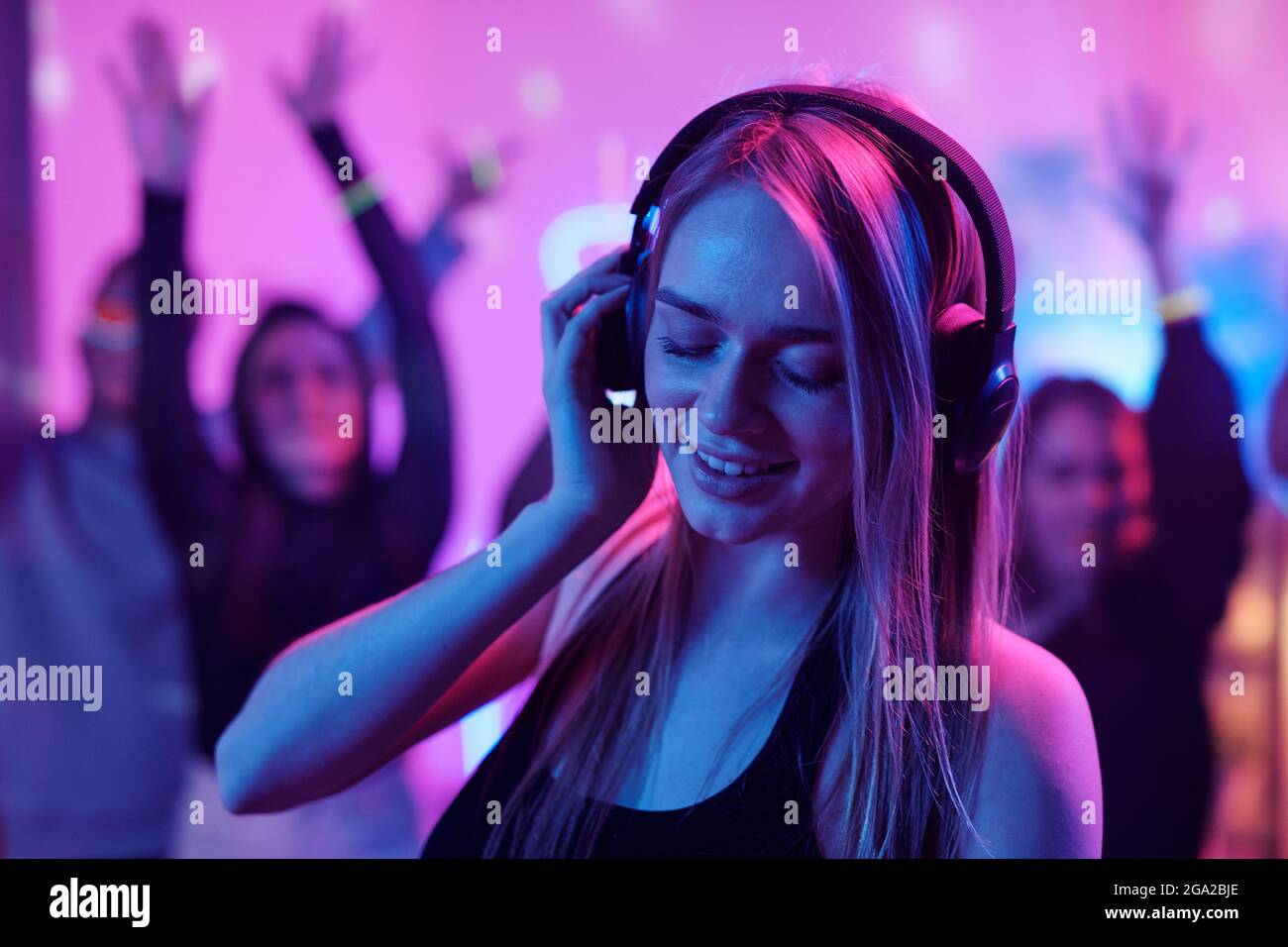 Jolie fille aux cheveux longs blonds touchant les écouteurs pendant le mixage des sons pour danser en discothèque et faire la fête avec des amis enthousiastes en arrière-plan Banque D'Images