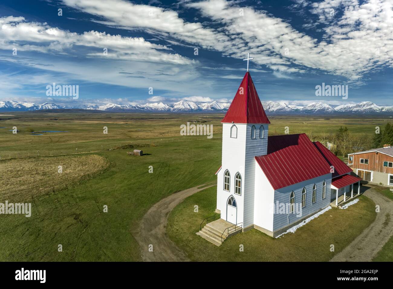 Vue aérienne de l'église au toit rouge, dans une prairie avec des nuages spectaculaires dans un ciel bleu et une chaîne de montagnes enneigée en arrière-plan, nous... Banque D'Images
