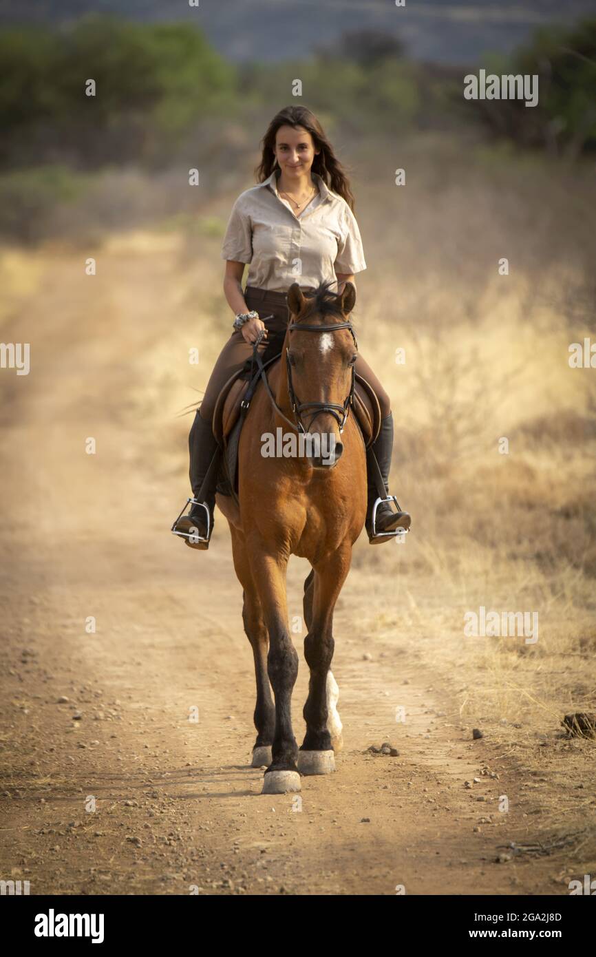 Femme à cheval (Equus ferus cabalus) sur une route de terre à travers le Bush sur la savane regardant la caméra et souriant au Gabus Game Ranch Banque D'Images