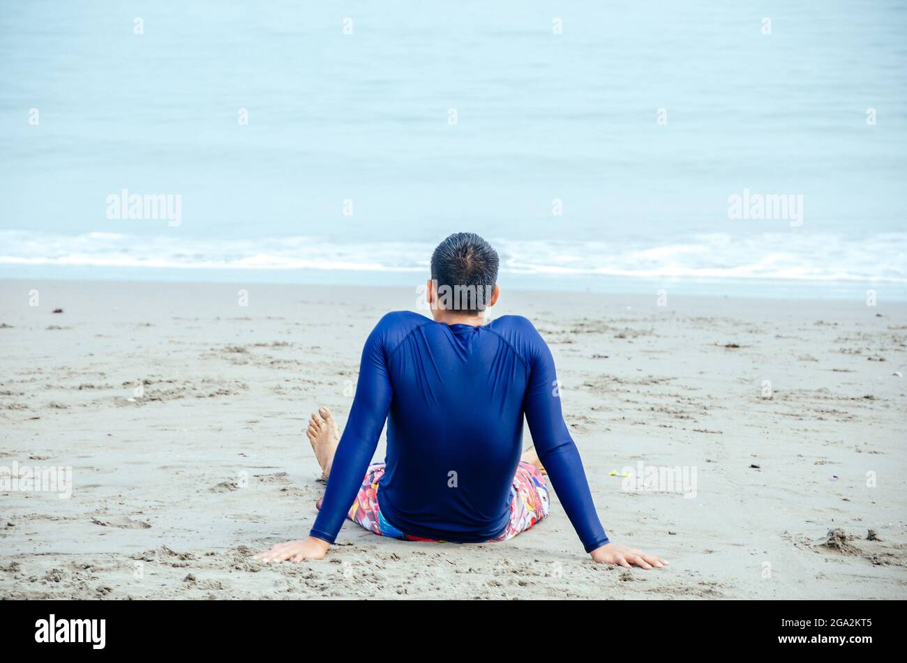 Jeune homme assis sur le sable dans un maillot de bain, face à la vague, méditant, pensant. Banque D'Images