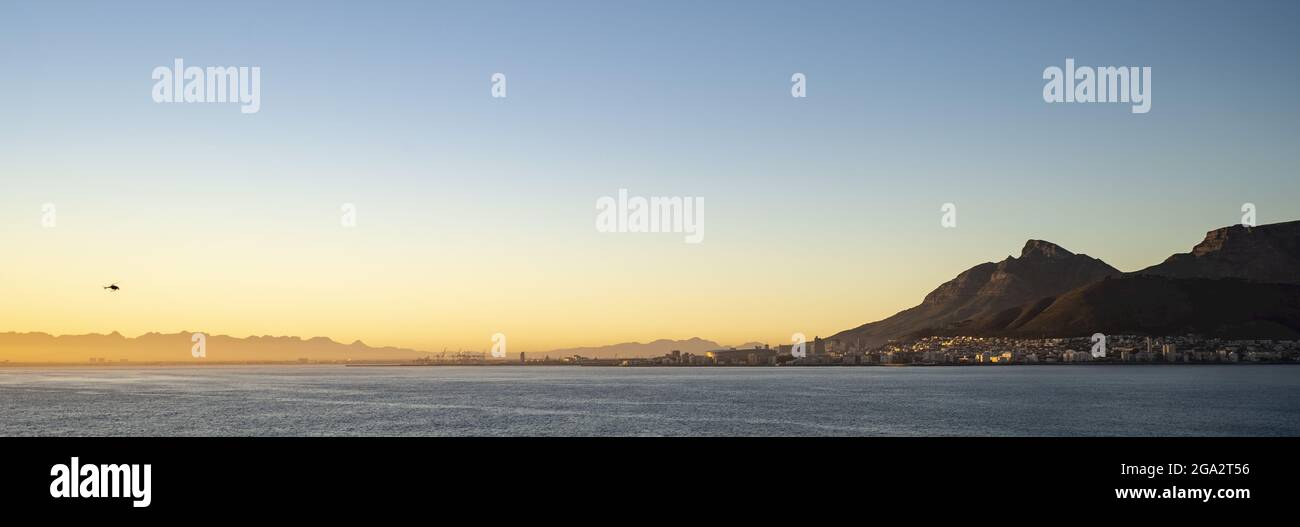 Silhouette d'un hélicoptère survolant Table Bay vers le port côtier et le port du Cap avec Devil's Peak et Table Mountain... Banque D'Images