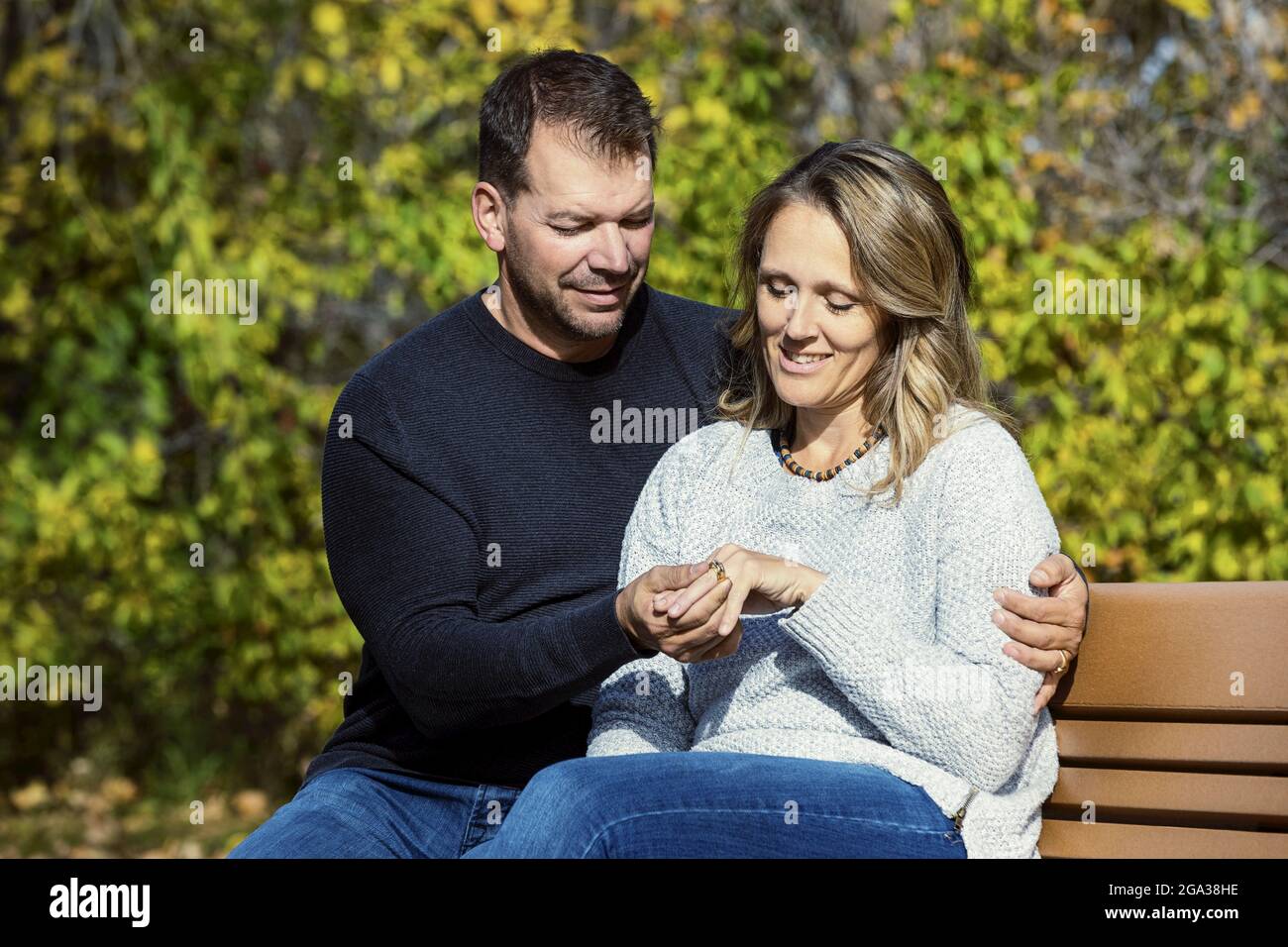 Un couple marié passant du temps de qualité ensemble à l'extérieur dans un parc de la ville pendant une chaude après-midi d'automne, regardant son anneau de mariage comme ils s'assoient sur un ... Banque D'Images