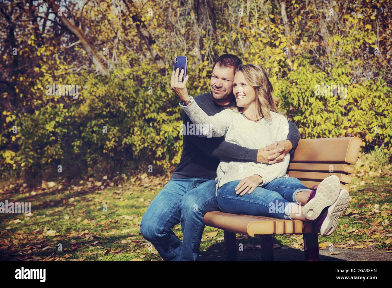 Un couple marié passant du temps ensemble de qualité à l'extérieur dans un parc de la ville pendant un après-midi d'automne chaud, prenant un autoportrait tout en étant assis sur une... Banque D'Images