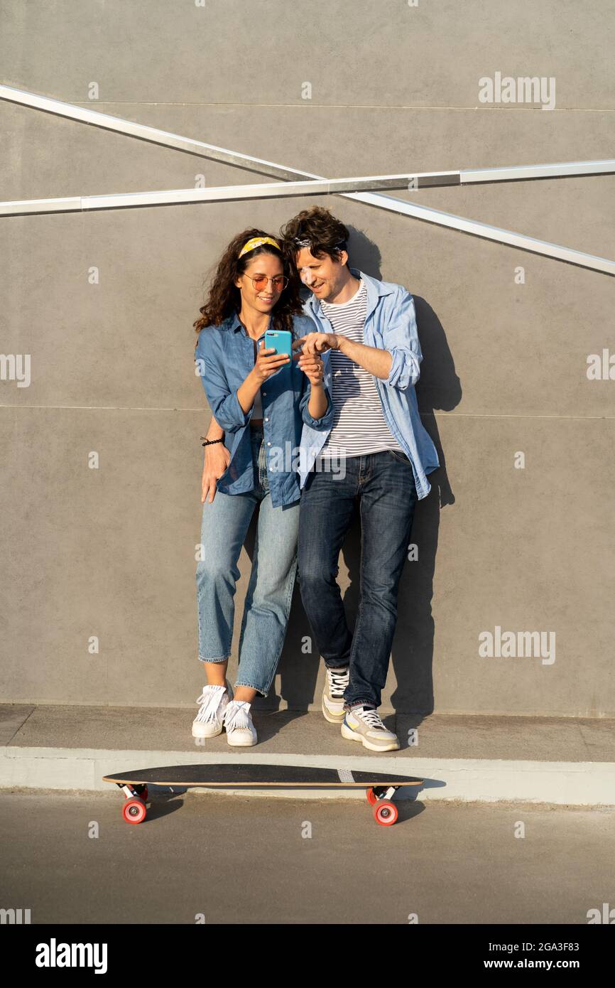 Jeune couple cool regardant sur l'écran de smartphone en plein air embrasser stand à la longue skate sourire heureux Banque D'Images