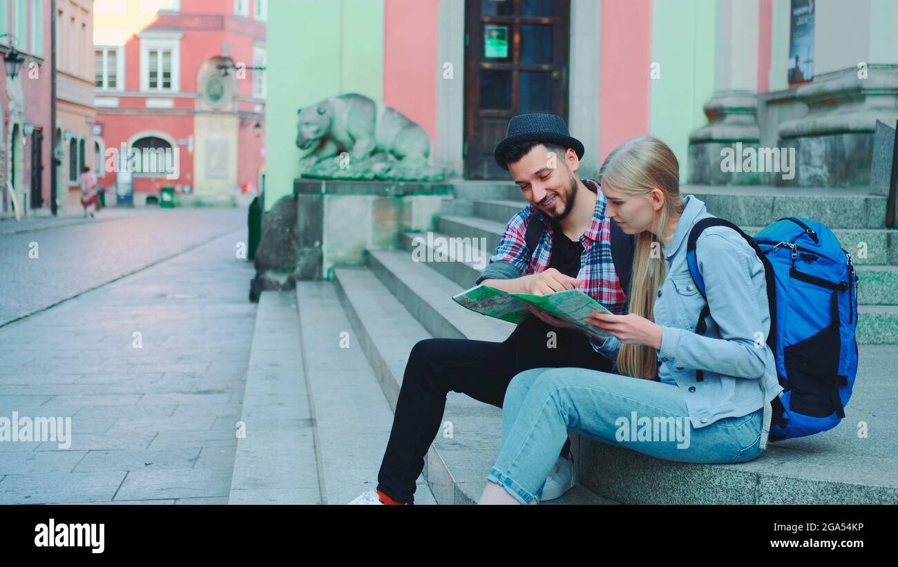 Joyeux couple de touristes assis dans les escaliers de la rue, en vérifiant le plan de la ville et en discutant. En arrière-plan, il y a une belle vue sur la vieille ville. Banque D'Images