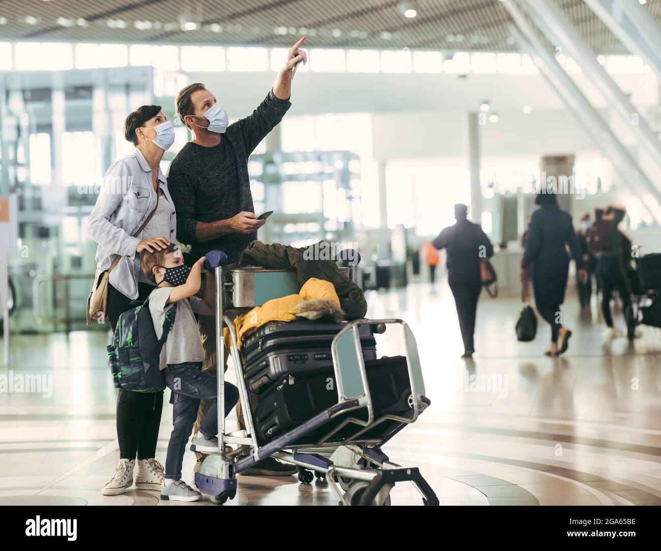 Homme pointant à l'arrivée à l'embarquement et femme parlante à l'aéroport. Famille avec un chariot à bagages attendant leur vol à l'aéroport. Banque D'Images