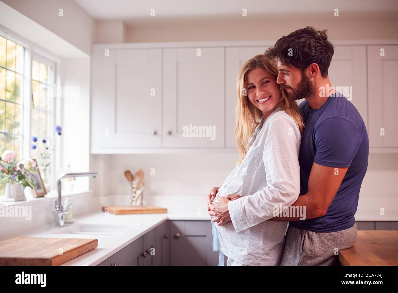 Couple avec homme embrassant femme enceinte portant un pyjama debout dans la cuisine tenant Bump Banque D'Images
