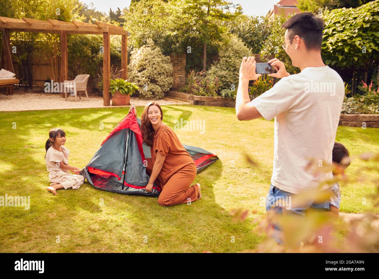 Père asiatique prenant des photos sur téléphone portable comme famille de tente d'installation dans le jardin à la maison Banque D'Images