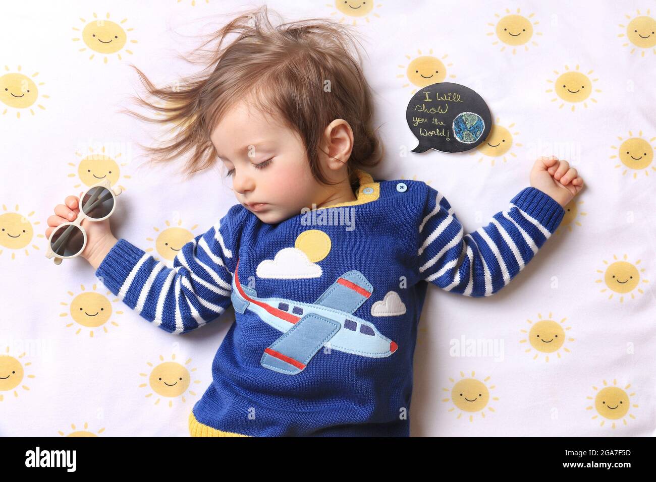 Un bébé endormi portant une chemise d'avion et tenant des lunettes de soleil rêvant de voyager, avec une bulle de parole disant que je vais vous montrer le monde. Banque D'Images