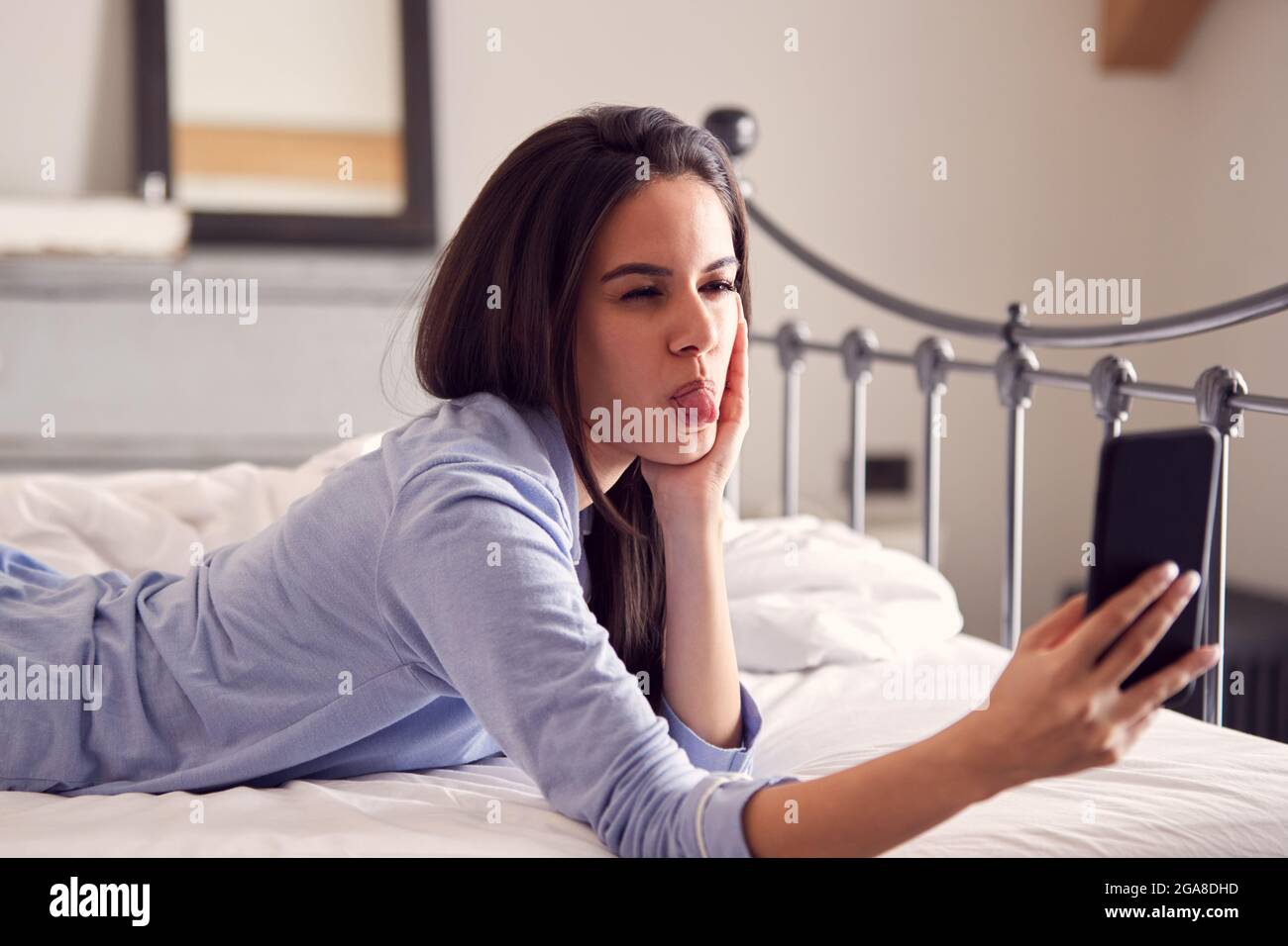 Femme portant un pyjama tirant sur le visage tout en prenant Selfie sur un téléphone portable couché sur le lit Banque D'Images