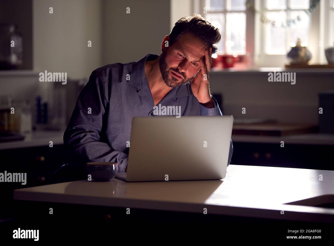 Malheureux homme portant un pyjama assis dans la cuisine la nuit avec un ordinateur portable Banque D'Images