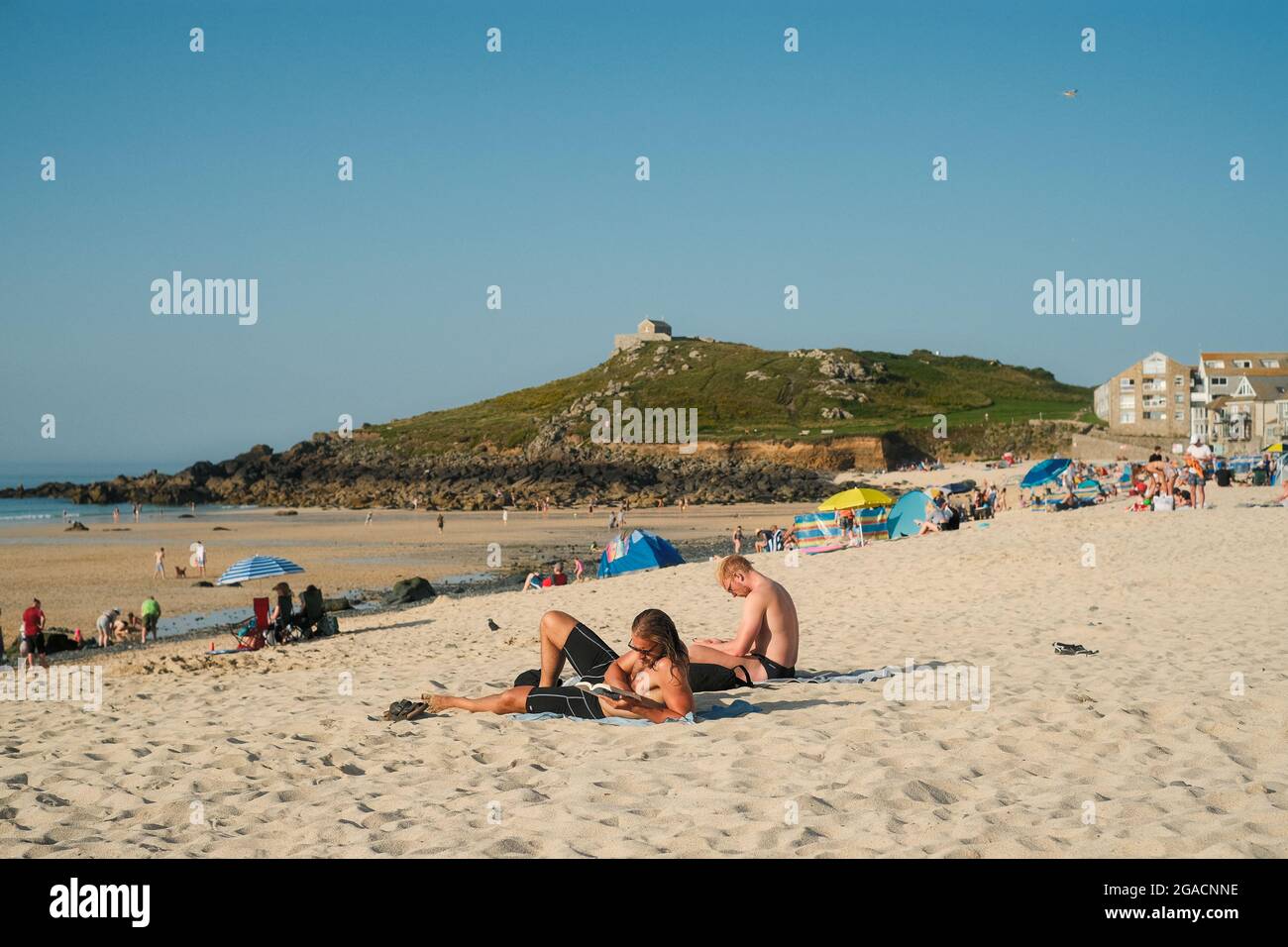 Deux jeunes hommes se sont assis à la lecture sur la plage de Porthmeor en été. St Ives, Cornwall, Royaume-Uni. Banque D'Images