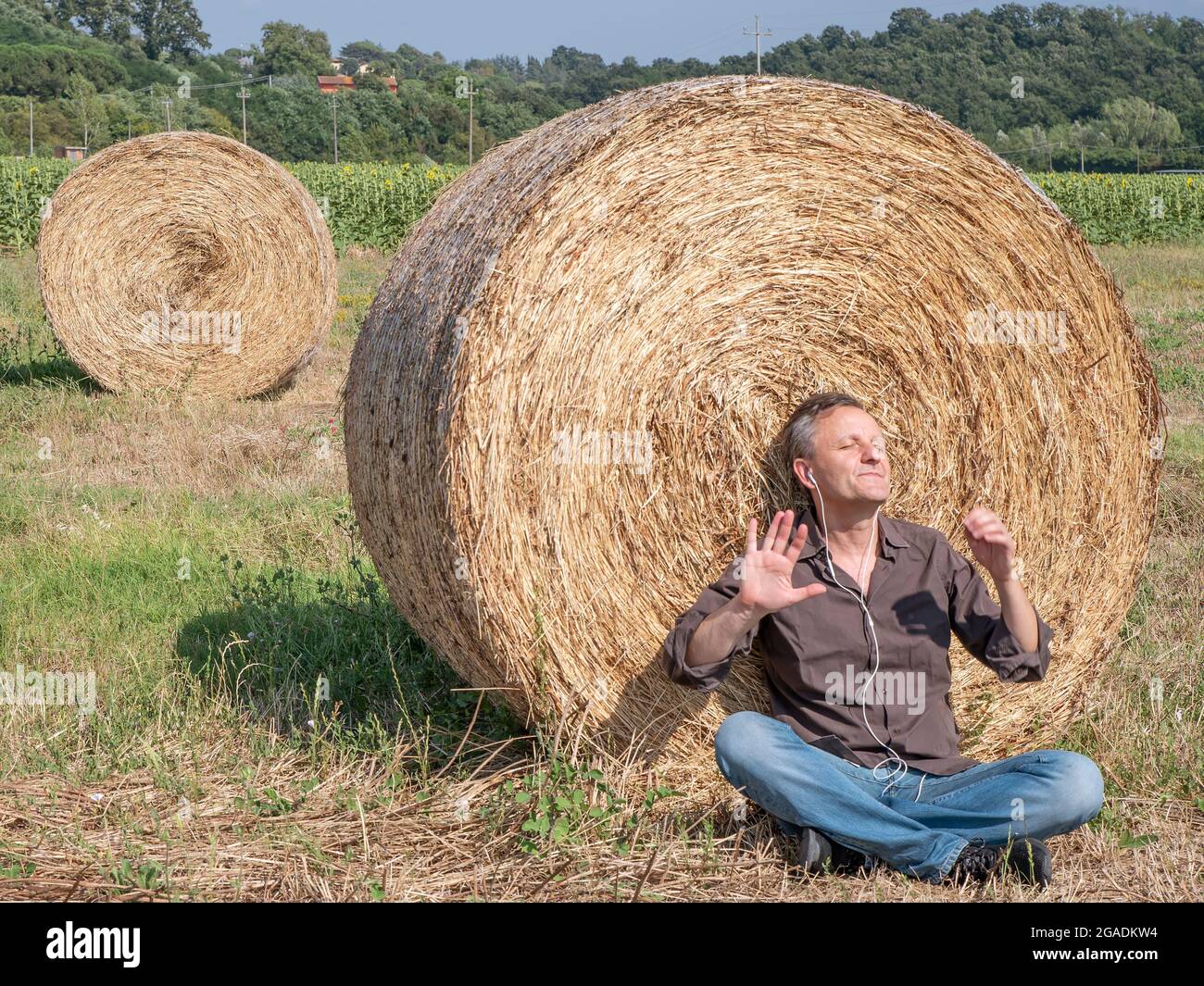 Un homme blanc d'âge moyen écoute de la musique sur des écouteurs assis dans un champ, avec son dos à une balle de foin Banque D'Images