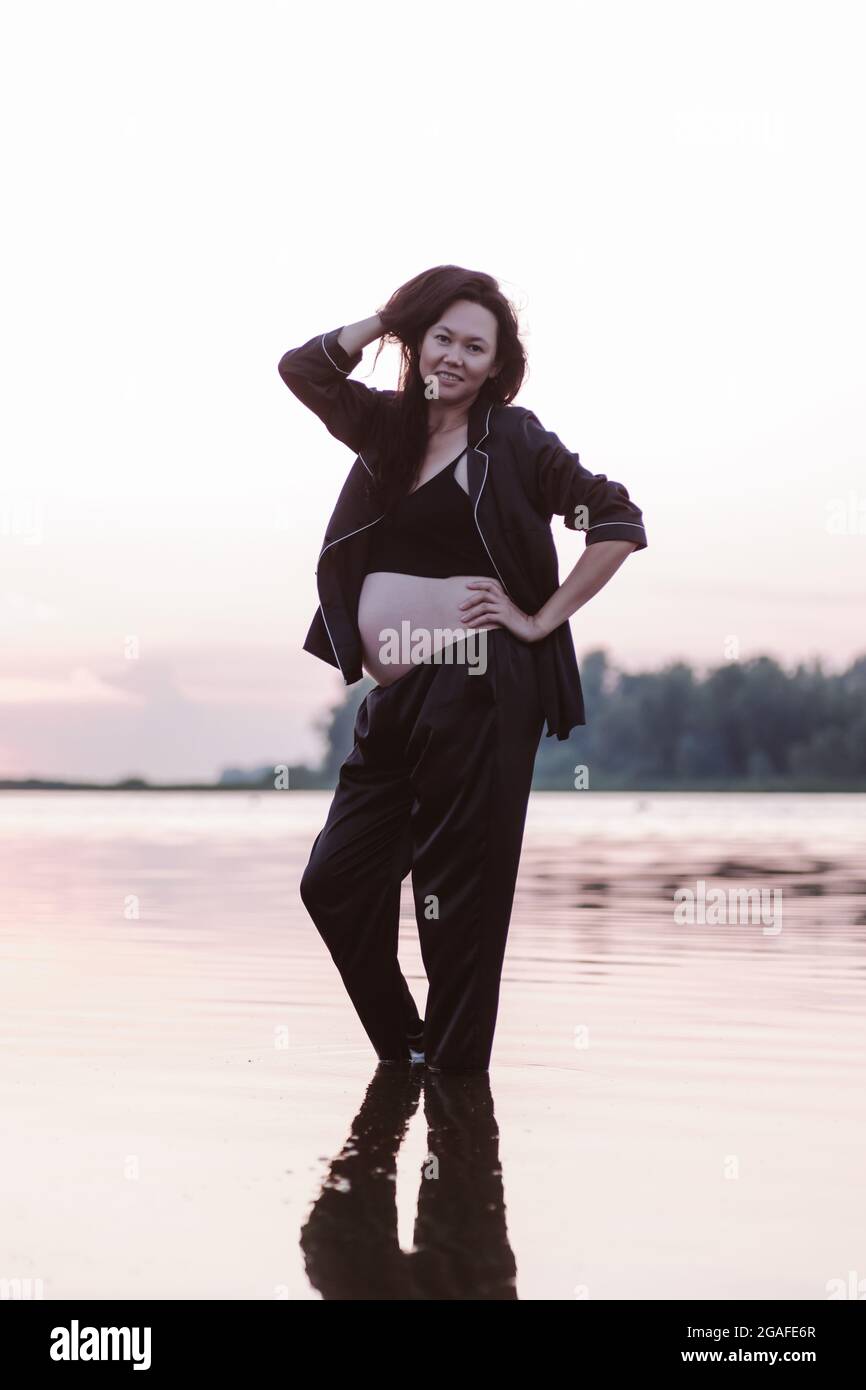 La femme pose dans l'eau. La jeune femme asiatique enceinte en costume sombre se tient dans l'eau sur la mer au coucher du soleil et tient ses longs cheveux bruns avec ses mains Banque D'Images