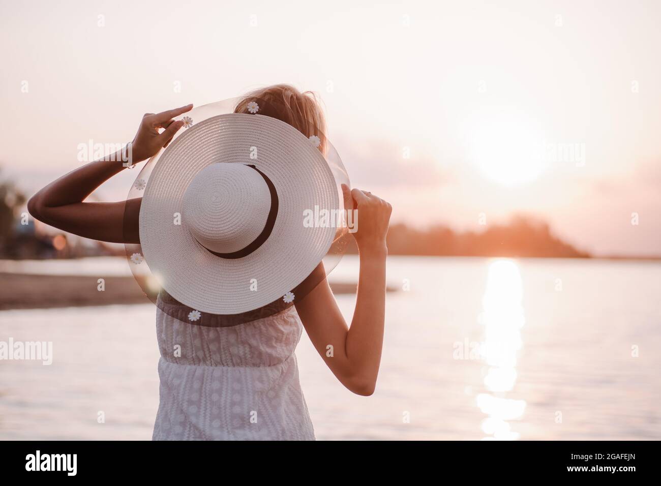 Une femme avec un chapeau au coucher du soleil vue arrière. Une jeune blonde dans une sunresson d'été et un chapeau de paille décoré d'un ruban et de fleurs pose contre le Banque D'Images