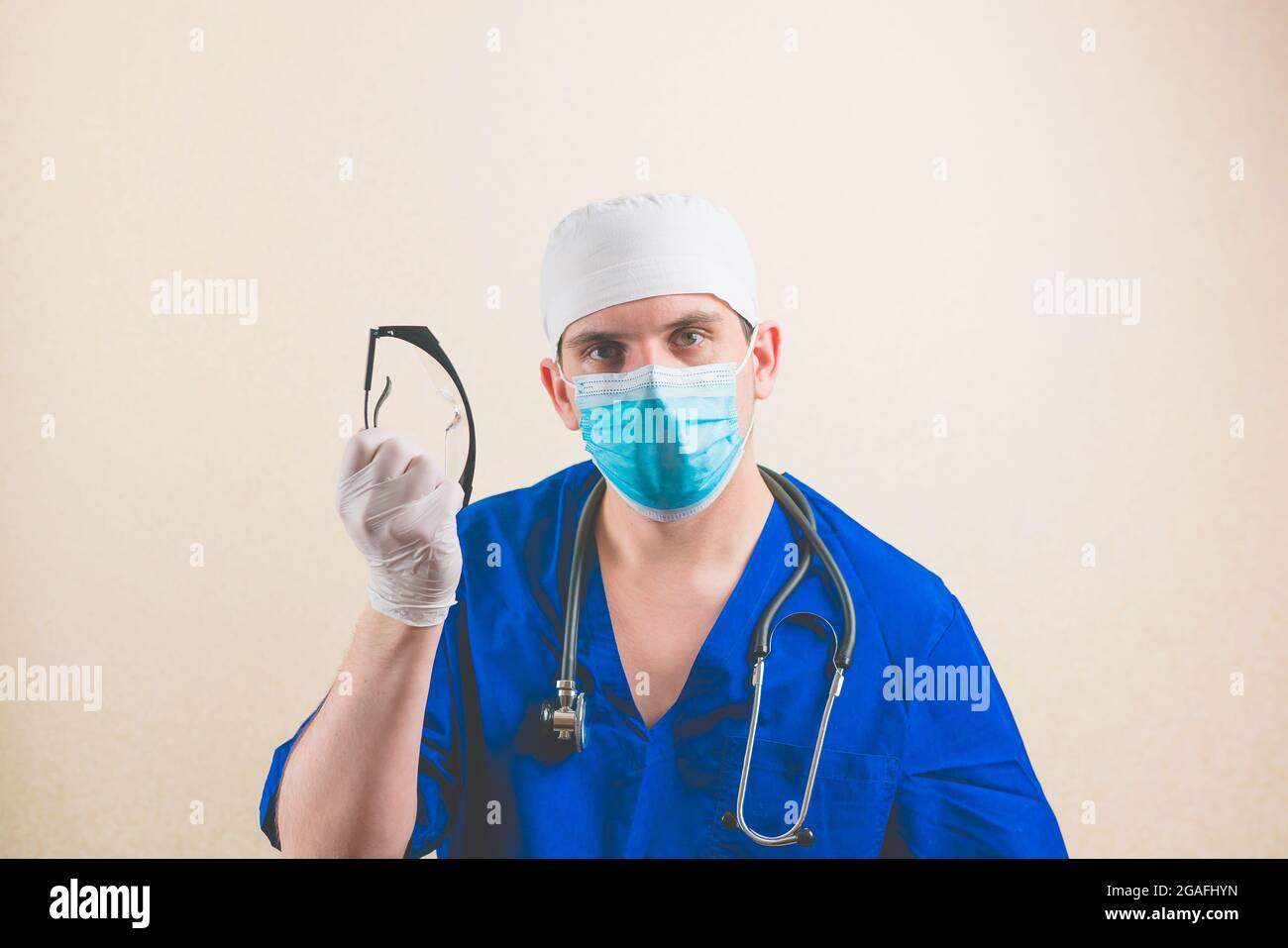 Jeune médecin en costume de chirurgien bleu et masque de protection respiratoire avec des lunettes de protection dans sa main isolée Banque D'Images