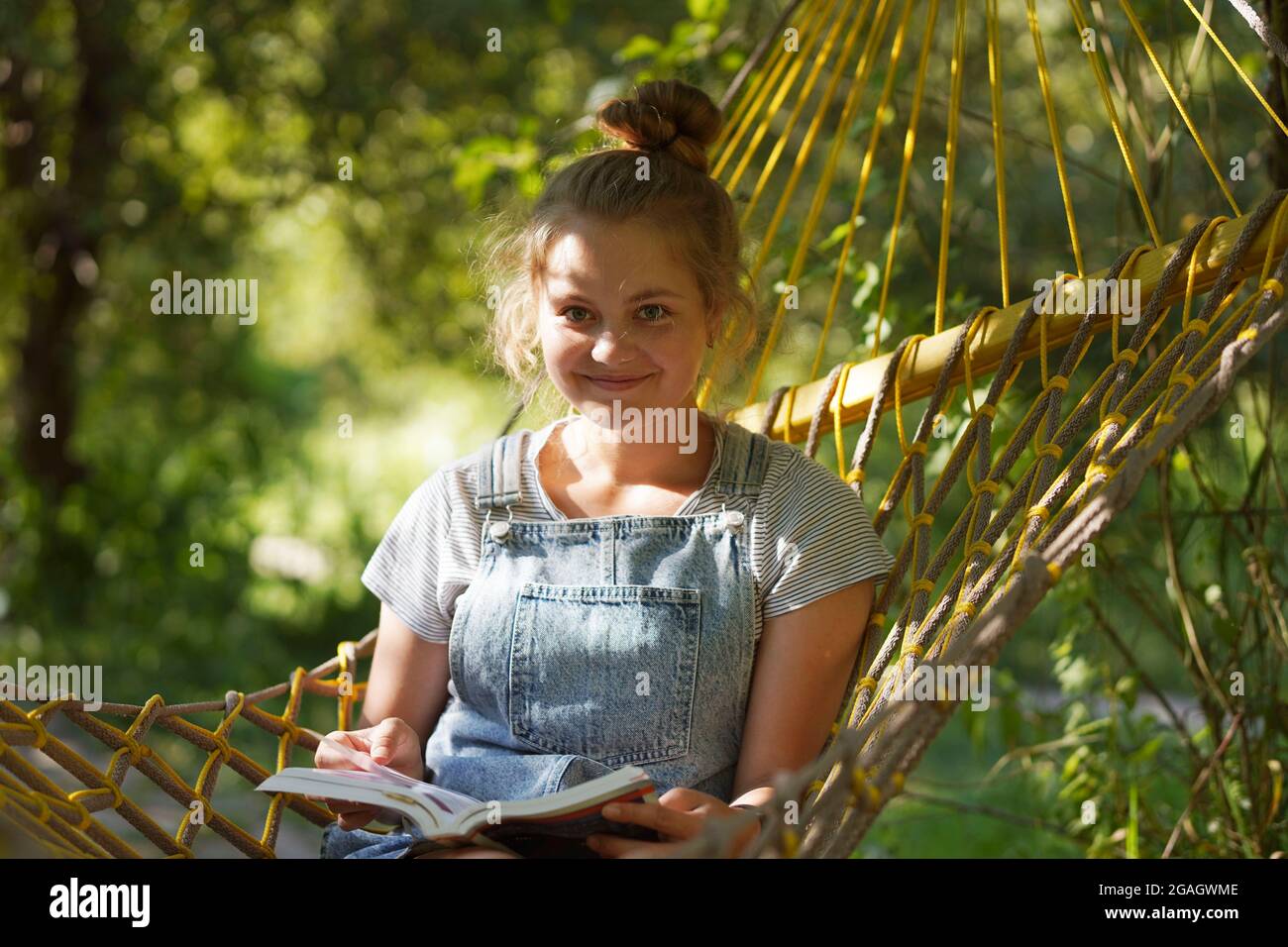 Jeune étudiante souriante lisant UN livre en étant allongé sur UN hamac dans le jardin. Femme se détendant dans un livre de lecture hamac. Retour à l'université, université. Banque D'Images