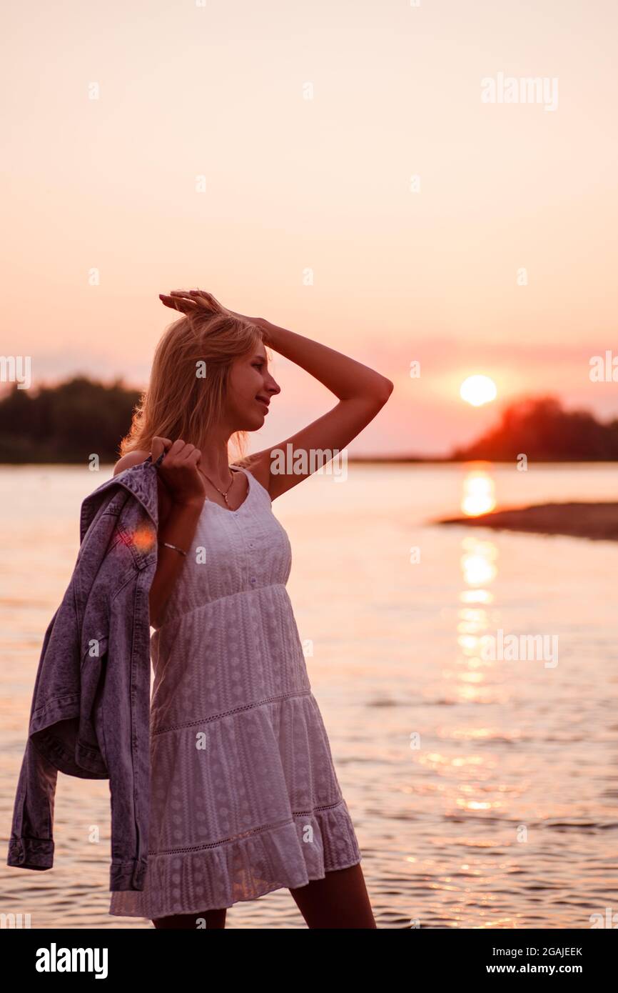 Photo dans le profil de la jeune femme. Une belle blonde heureuse en blanc  de la sundrress d'été se tient sur la rive de la rivière et tient une veste  en denim