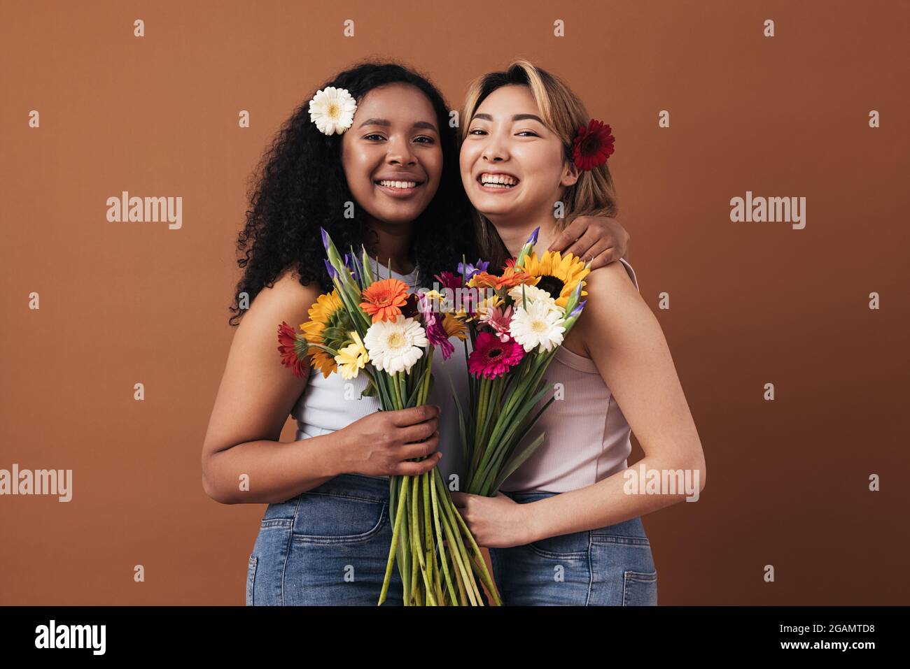 Deux jeunes femmes de différentes races avec des bouquets et des fleurs dans leurs cheveux posant sur un fond marron Banque D'Images