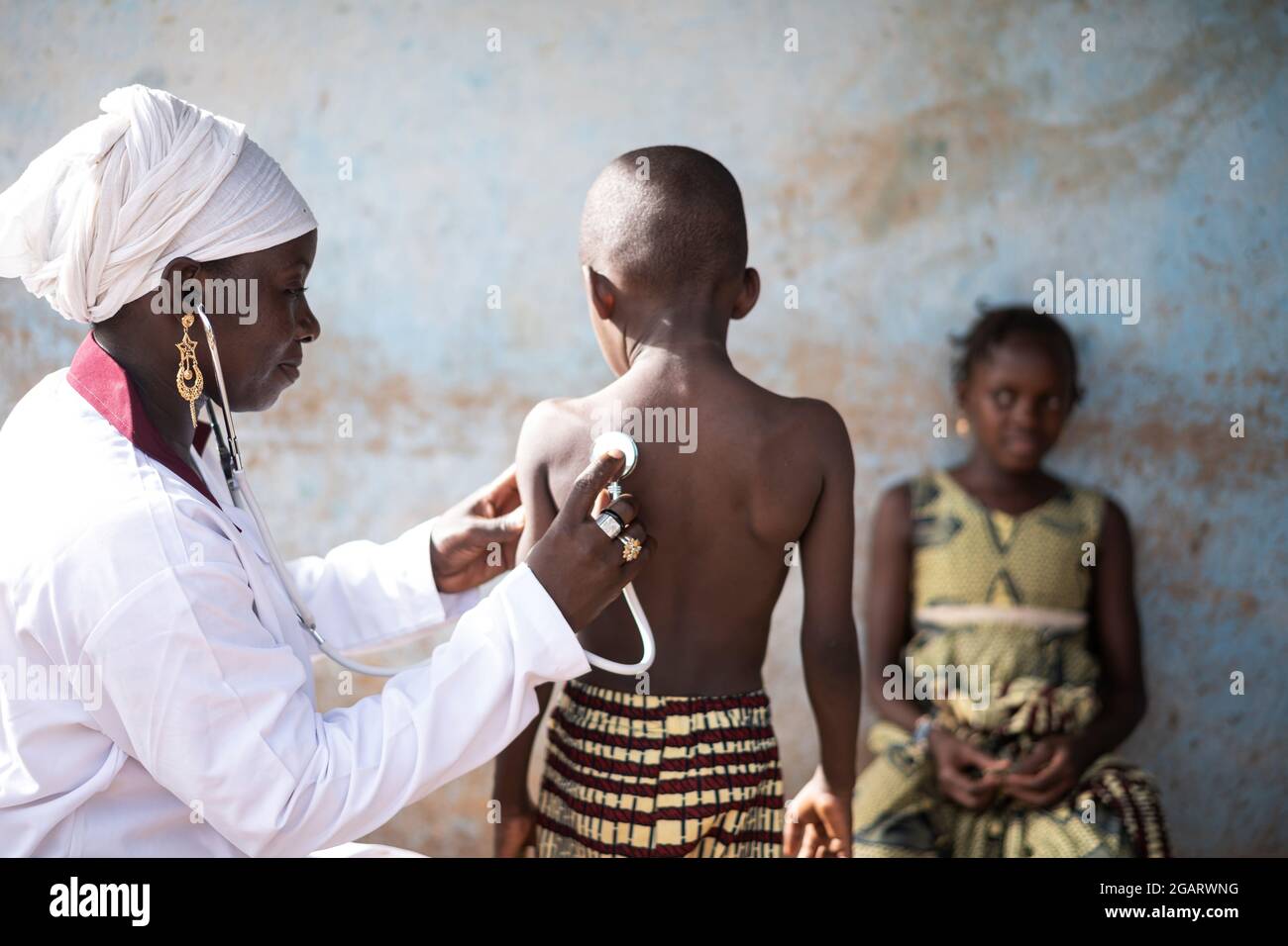 Dans cette image, un médecin noir souriant, assis dans une salle de classe, ausculte l'un des deux petits enfants africains avec un stéthoscope pendant l'école COVID Banque D'Images