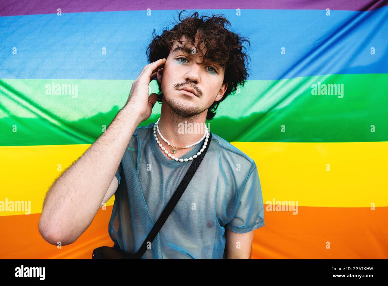 Homme gay embrassant son identité. Jeune gay regardant la caméra tout en se tenant contre un drapeau de fierté. Jeune homme queer portant des fards à paupières contre le LGB Banque D'Images