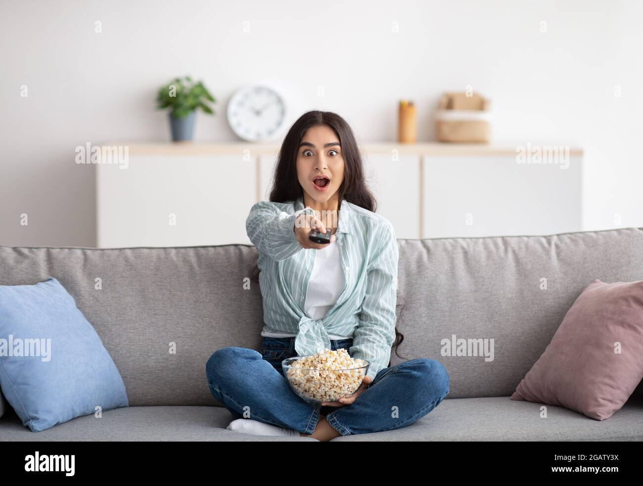 Pleine longueur de femme indienne choquée assis sur un canapé, manger du pop-corn, changer de chaîne, regarder la télévision à la maison. Une jeune femme surprise avec télécommande Banque D'Images