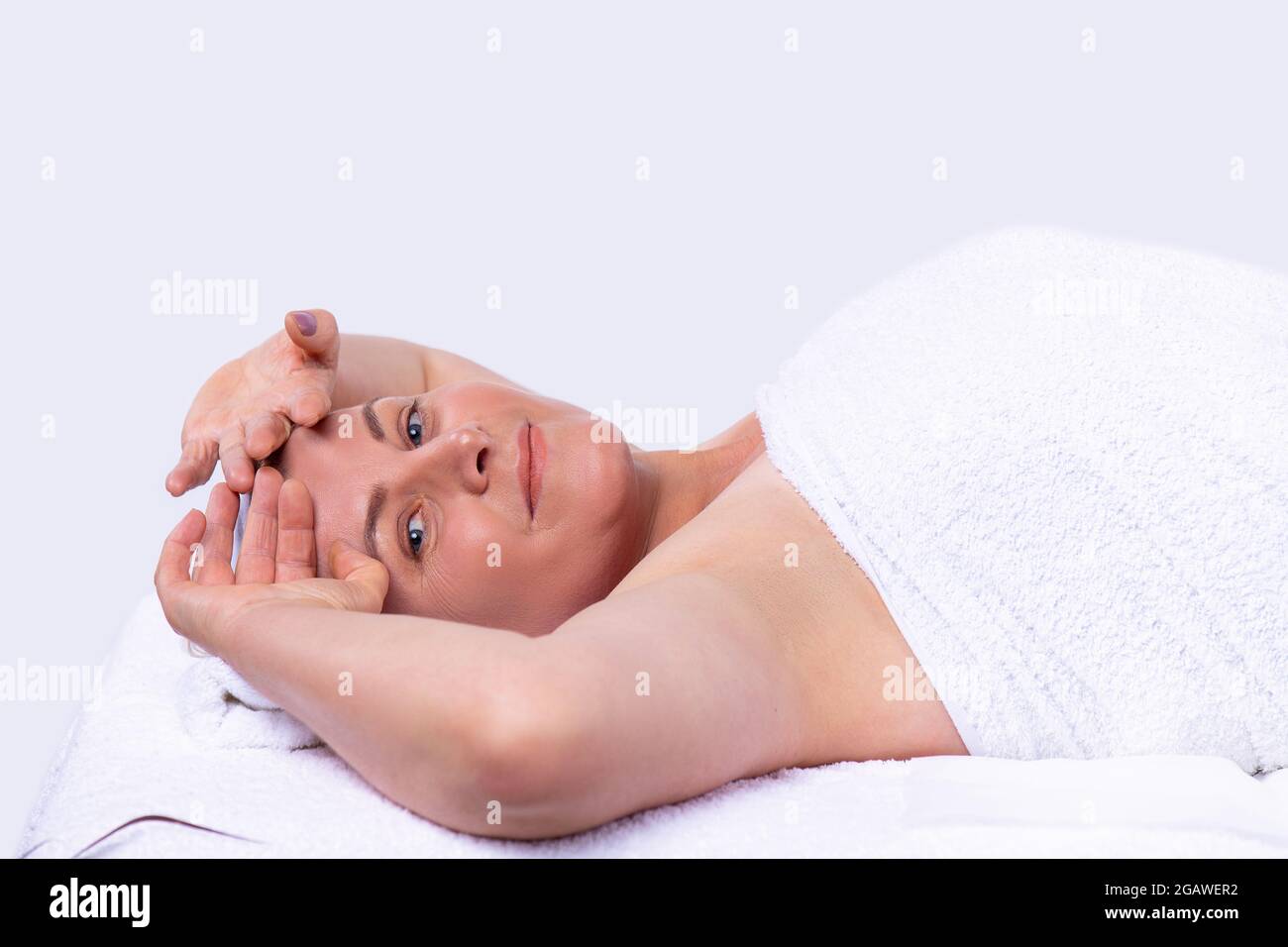 Femme calme et détendue allongé sur la table de massage dans une serviette blanche en gardant les mains sur son front en attendant les procédures de massage du visage. Banque D'Images