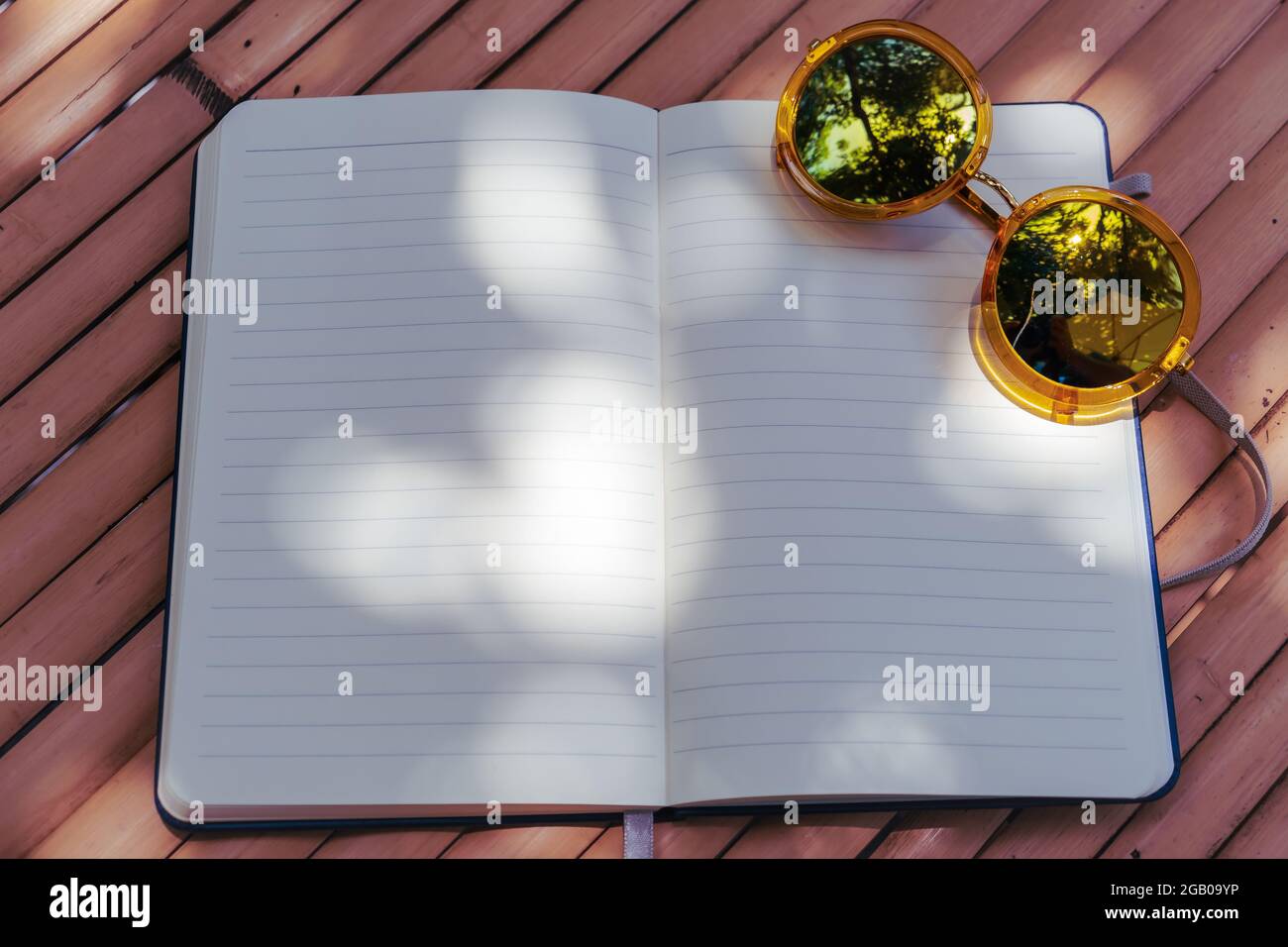 Ligne vide carnet écriture à la main avec lunettes de soleil flous sur une table en bambou en bois à l'espace de travail extérieur jardin décoré avec l'ombre et la lumière du soleil f Banque D'Images