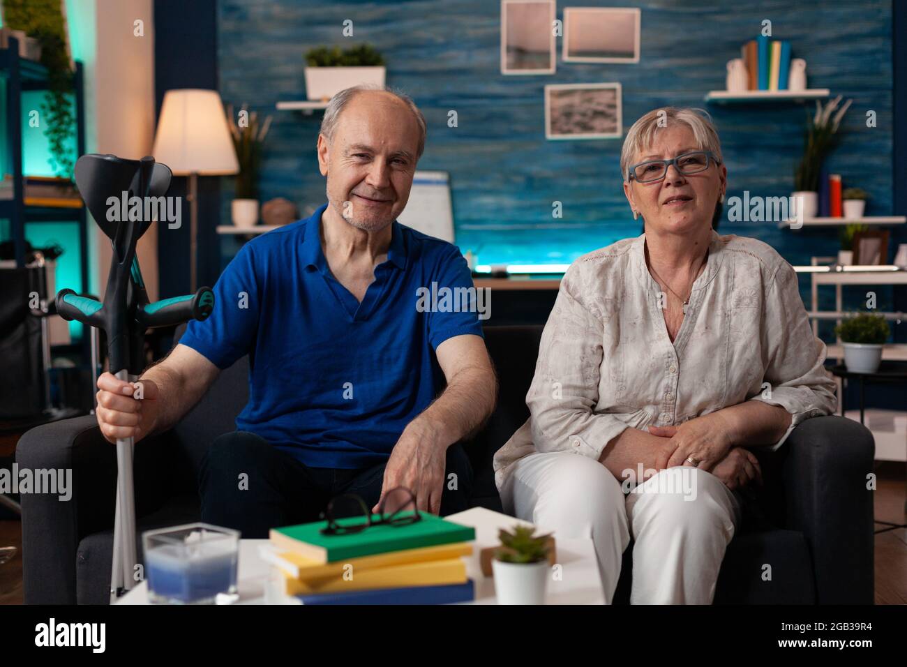 Portrait d'un couple âgé assis ensemble à la maison dans un salon décoré. Un vieux mari caucasien avec une béquille de transport pour personnes handicapées et une femme âgée souriant regardant la caméra Banque D'Images