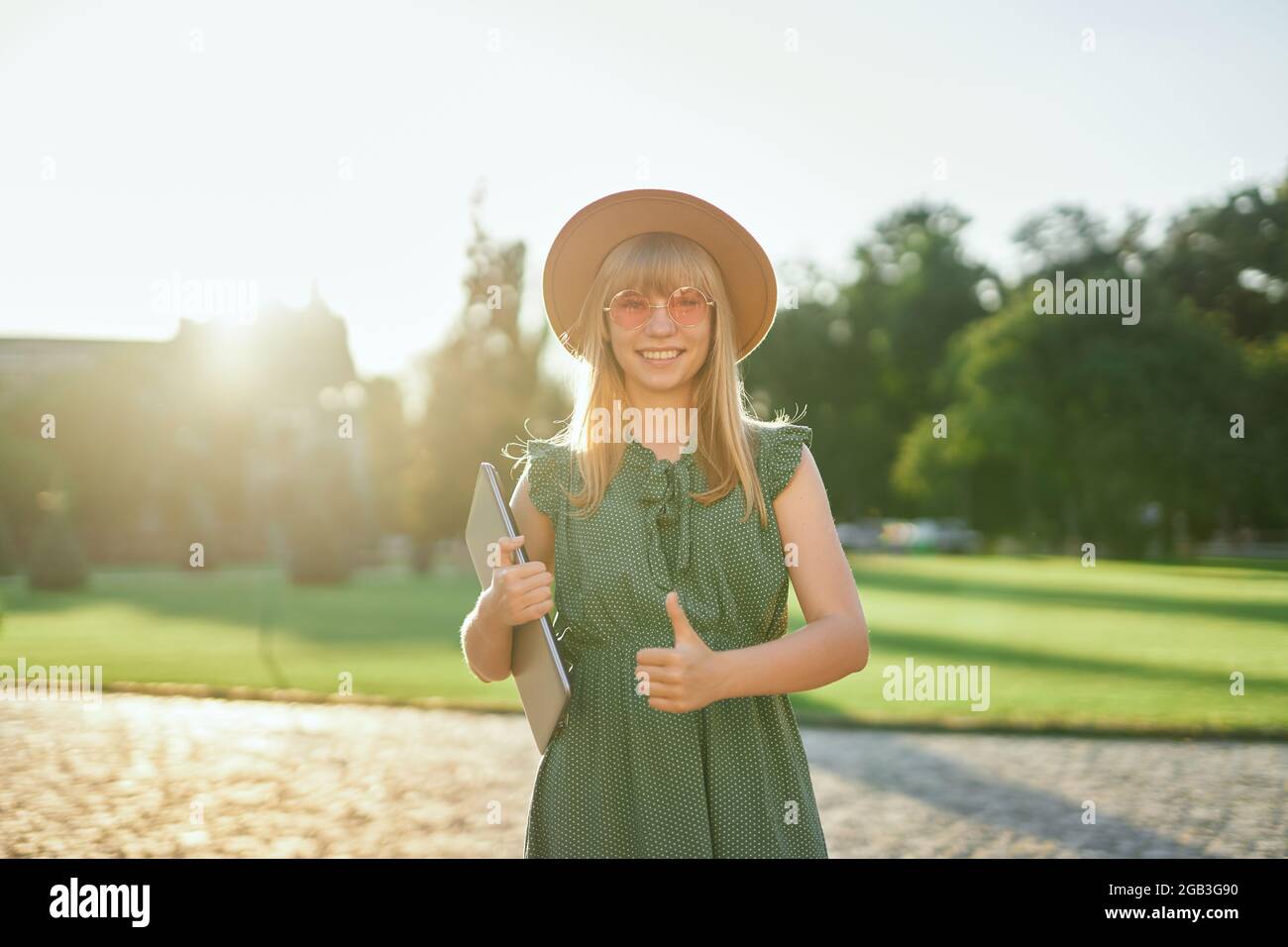 Jeune femme blonde et gaie étudiante à l'université ou à l'université avec  un ordinateur portable portant une robe verte et un chapeau sur le campus  de l'université montrant le pouce vers le