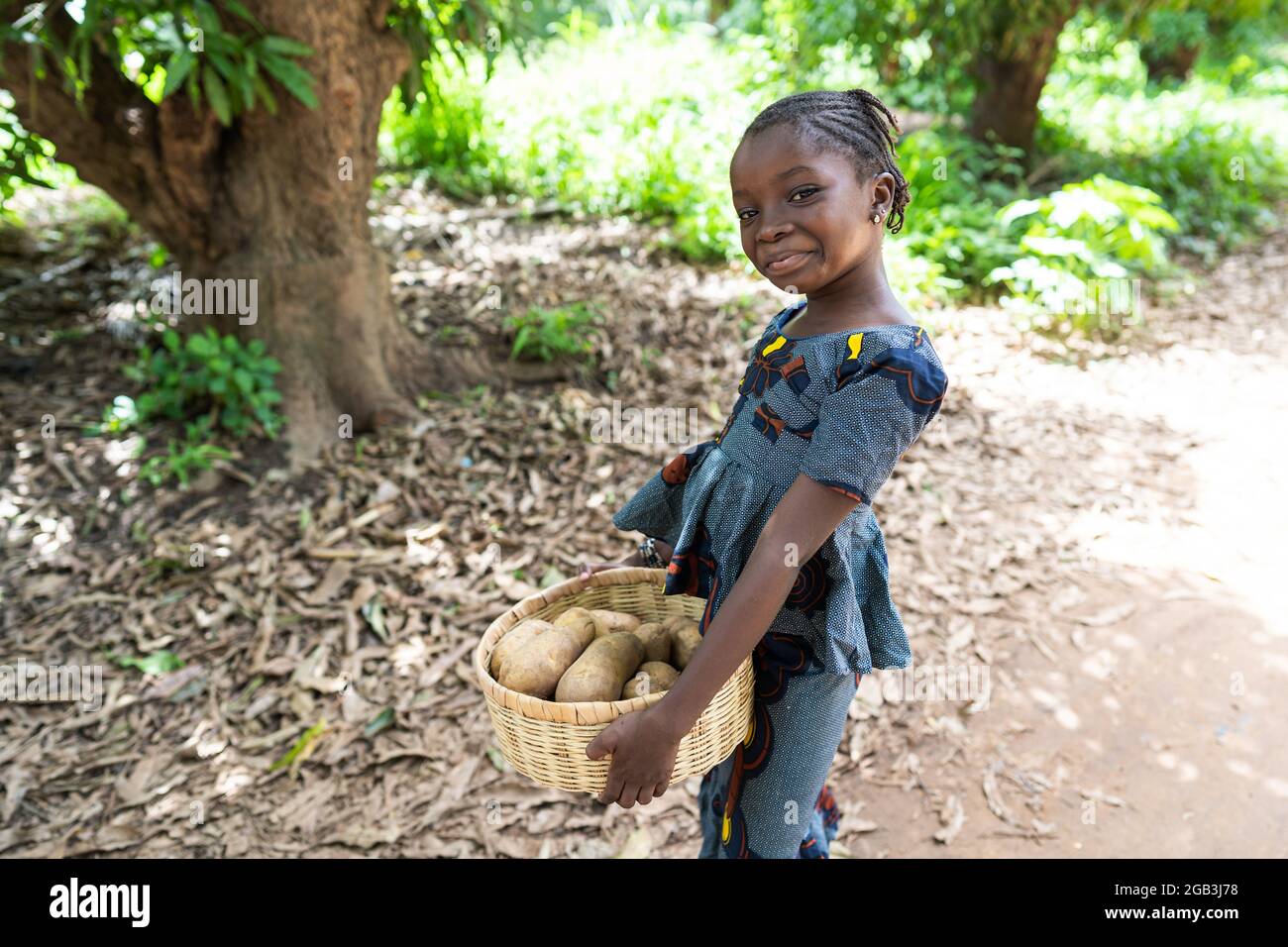 Dans cette image, une petite fille noire grinçant sur son chemin à la maison porte un panier de pommes de terre achetées sur le marché local Banque D'Images