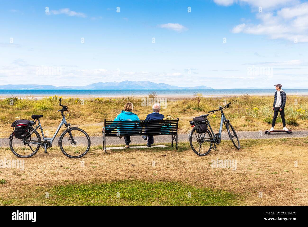 Deux cyclistes âgés assis sur un banc avec leurs cycles à côté d'eux comme un jeune homme sur un skateboard court près, surplombant le Firth de Clyde vers t Banque D'Images