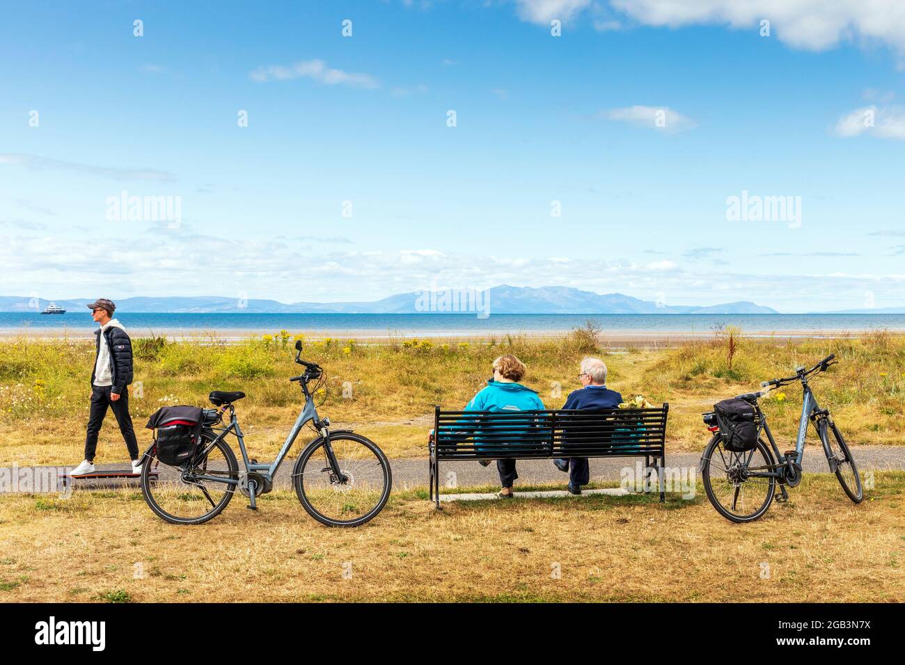 Deux cyclistes âgés assis sur un banc avec leurs cycles à côté d'eux comme un jeune homme sur un skateboard court près, surplombant le Firth de Clyde vers t Banque D'Images