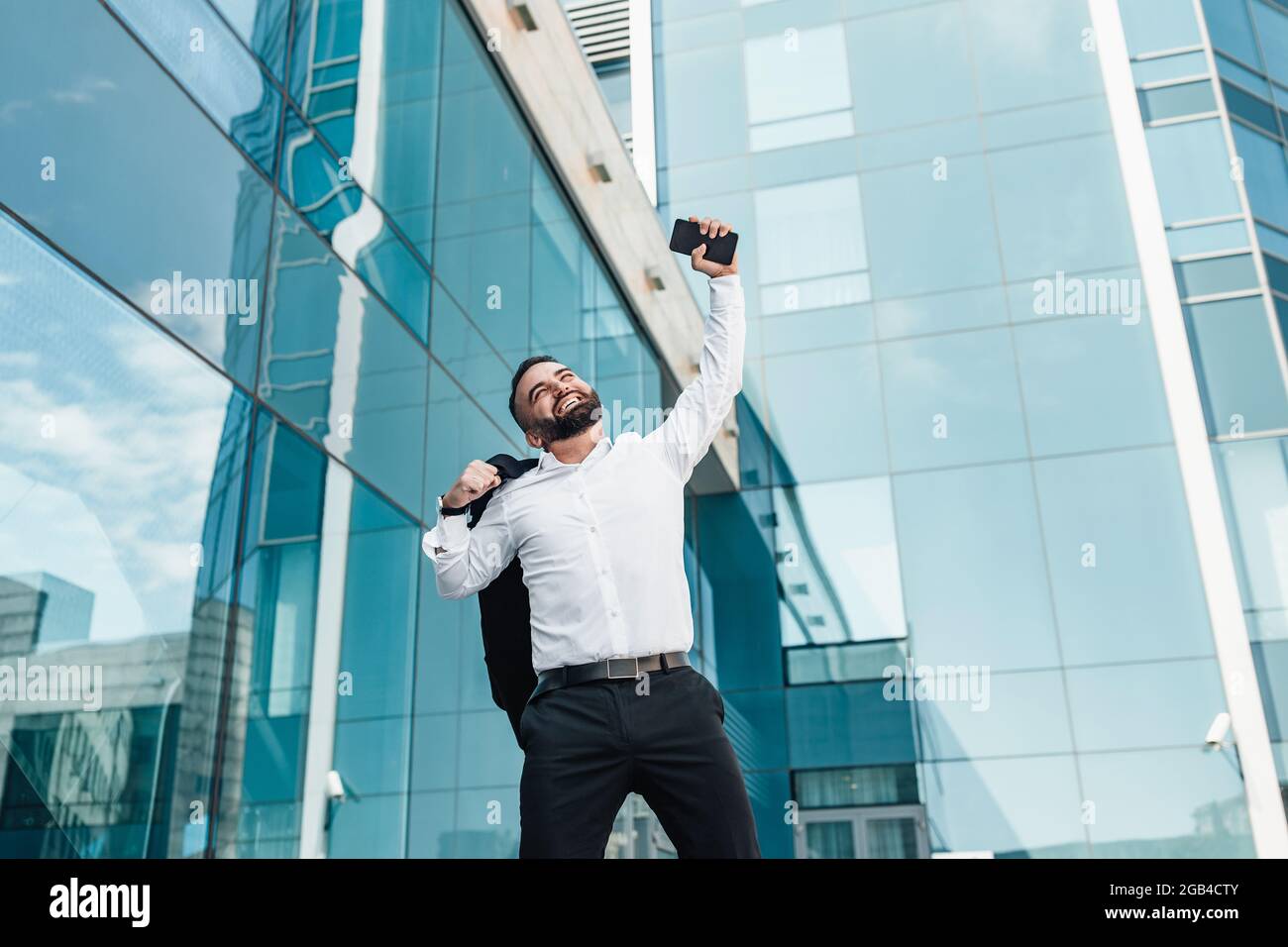 Homme d'affaires ecstatique mature célébrant le succès et levant la main vers le haut, debout à l'extérieur près du centre de bureau, espace vide Banque D'Images