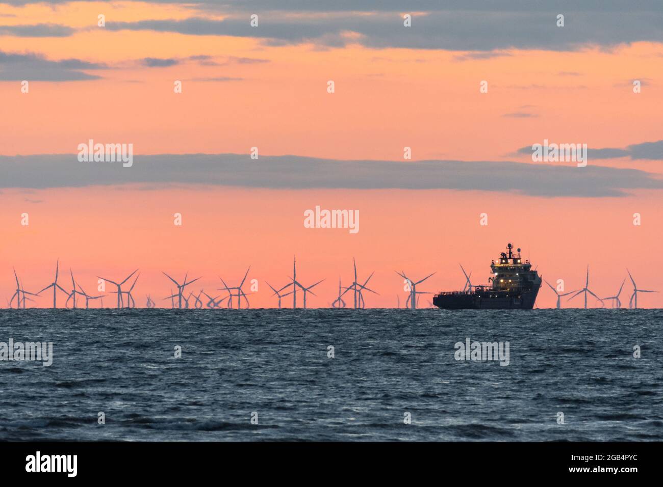 Ferme éolienne dans la mer d'Irlande au crépuscule. Un effet mirage fait que les turbines à l'horizon semblent fléchir lorsqu'elles tournent. Un navire d'approvisionnement est amarré Banque D'Images