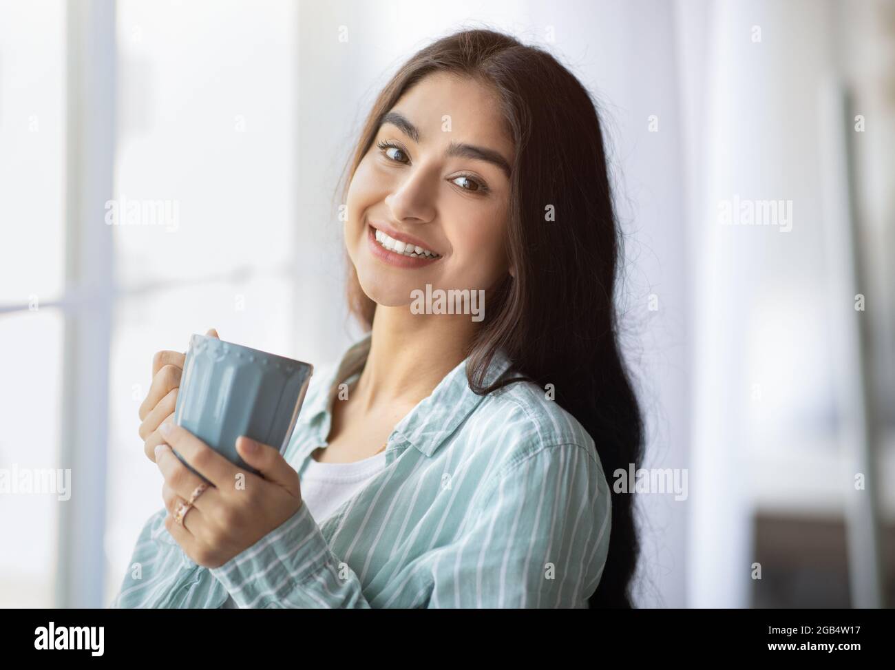 Portrait d'une jolie dame indienne debout près de la fenêtre avec une tasse de boisson chaude, souriant à l'appareil photo, à l'intérieur Banque D'Images