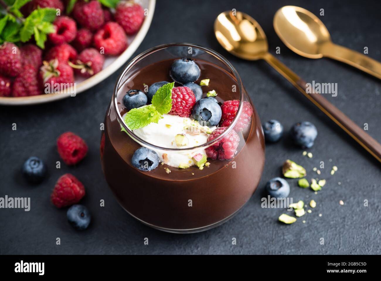 Mousse au chocolat noir avec pistaches, baies et crème en pot, fond de pierre noire, vue rapprochée Banque D'Images