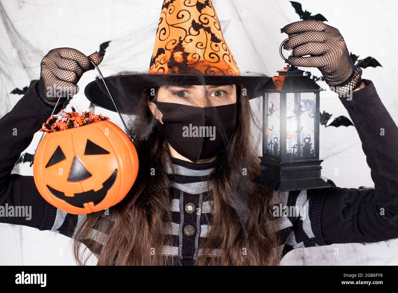 Halloween personnes masquées. Une femme en costume de sorcière dans un masque de protection tient une citrouille avec des bonbons et une lanterne lumineuse. Au cours du cycle de coronavirus Banque D'Images