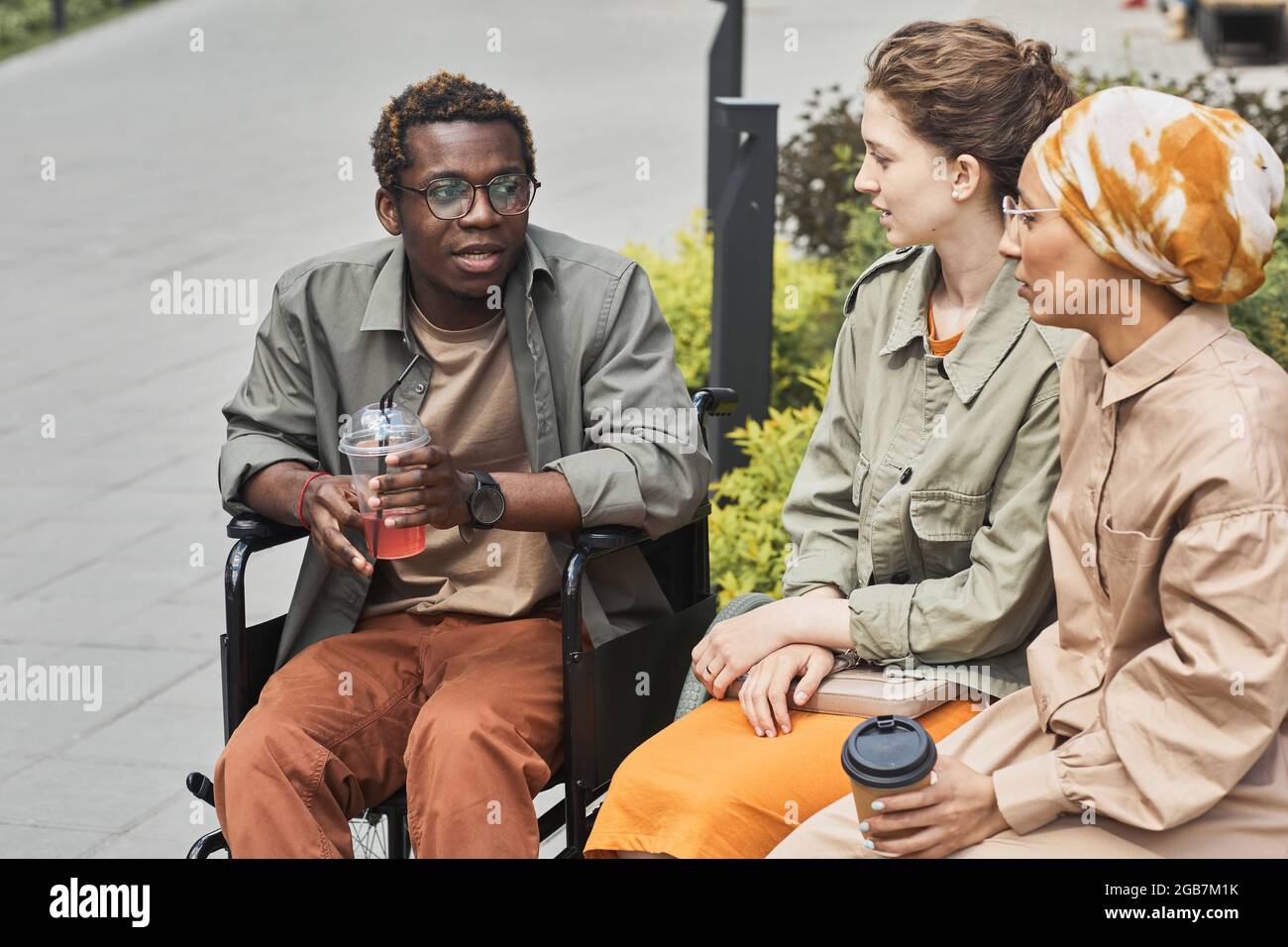 Jeune homme noir handicapé assis en fauteuil roulant et buvant de la limonade tout en partageant l'histoire avec des amis à l'extérieur Banque D'Images
