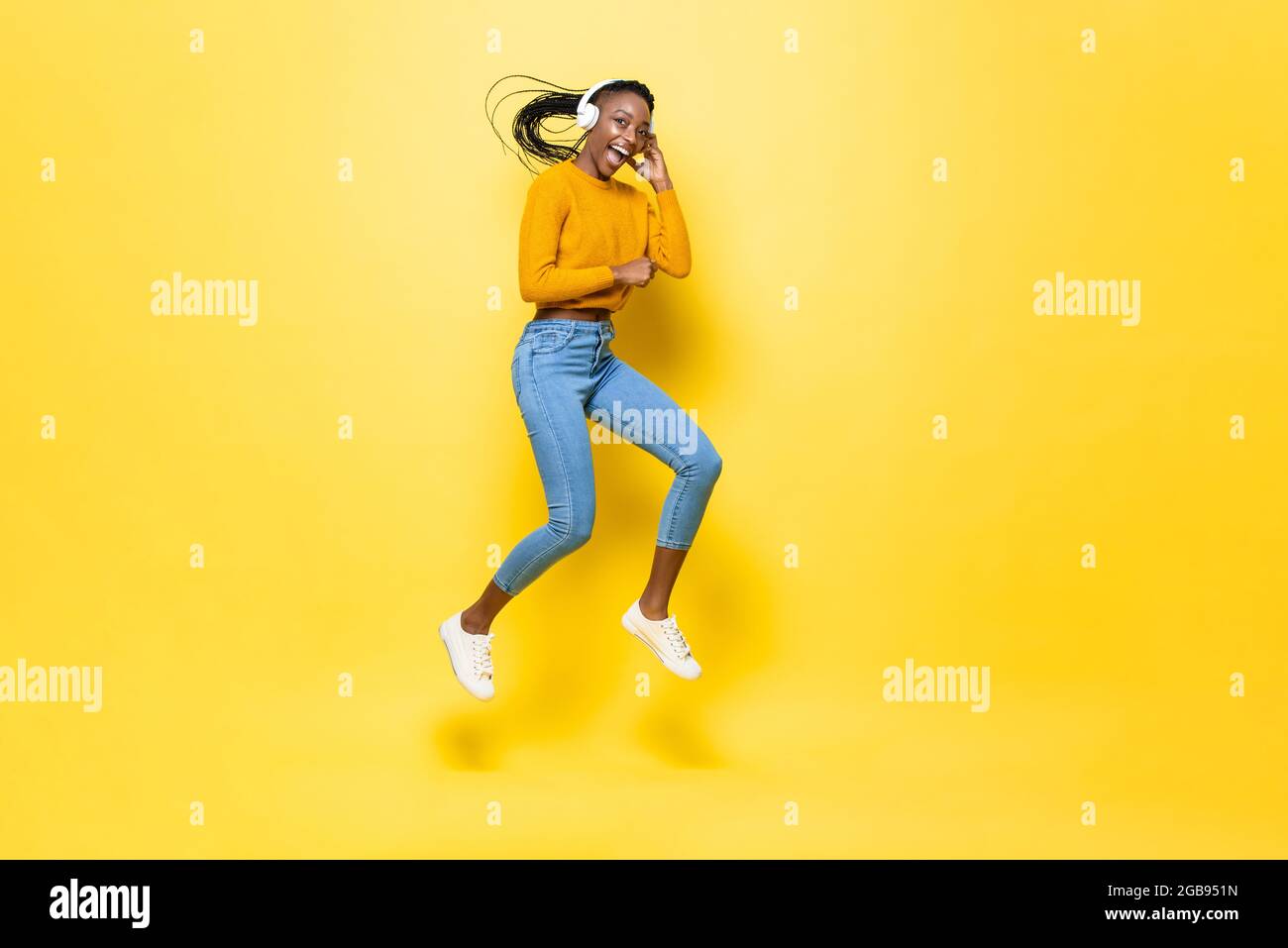 Bonne jeune femme afro-américaine énergique portant un casque à l'écoute de la musique et sautant dans un arrière-plan de studio isolé jaune Banque D'Images