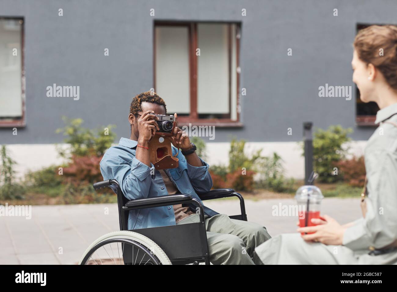 Photographe afro-américain assis en fauteuil roulant et utilisant un appareil photo traditionnel tout en prenant la photo d'une femme avec un verre Banque D'Images