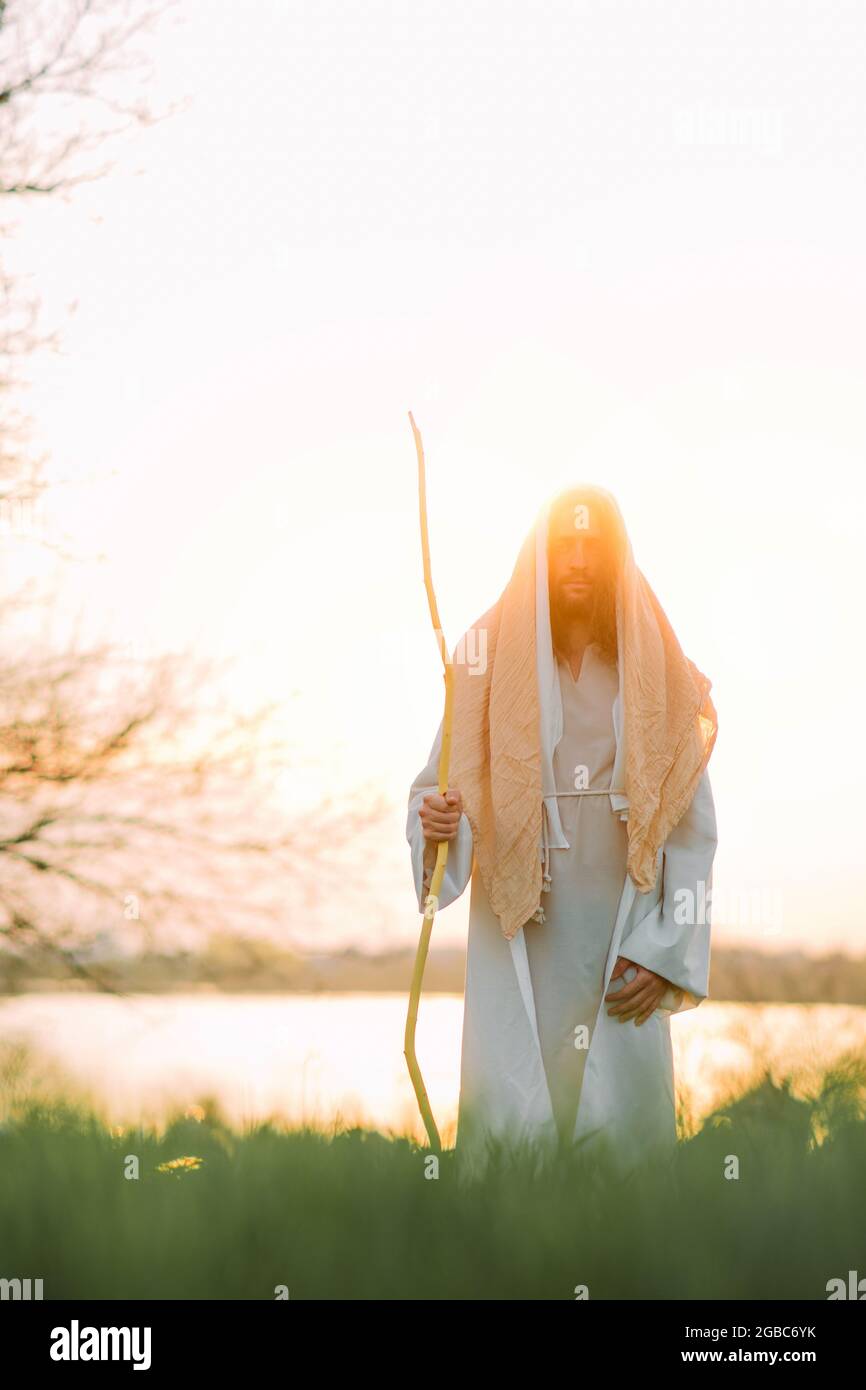 Jésus Christ avec un personnel en bois se tient dans un pré vêtu de sa robe blanche traditionnelle contre fond de rivière et de coucher de soleil. Banque D'Images