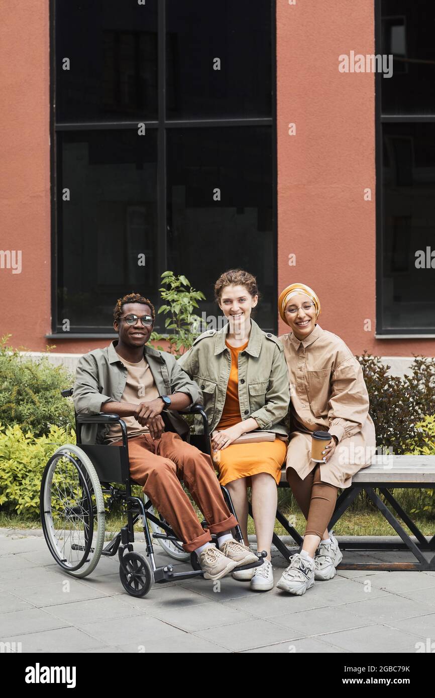 Portrait d'un jeune homme afro-américain souriant et handicapé assis en fauteuil roulant et posant avec des femmes multiethniques sur le banc Banque D'Images