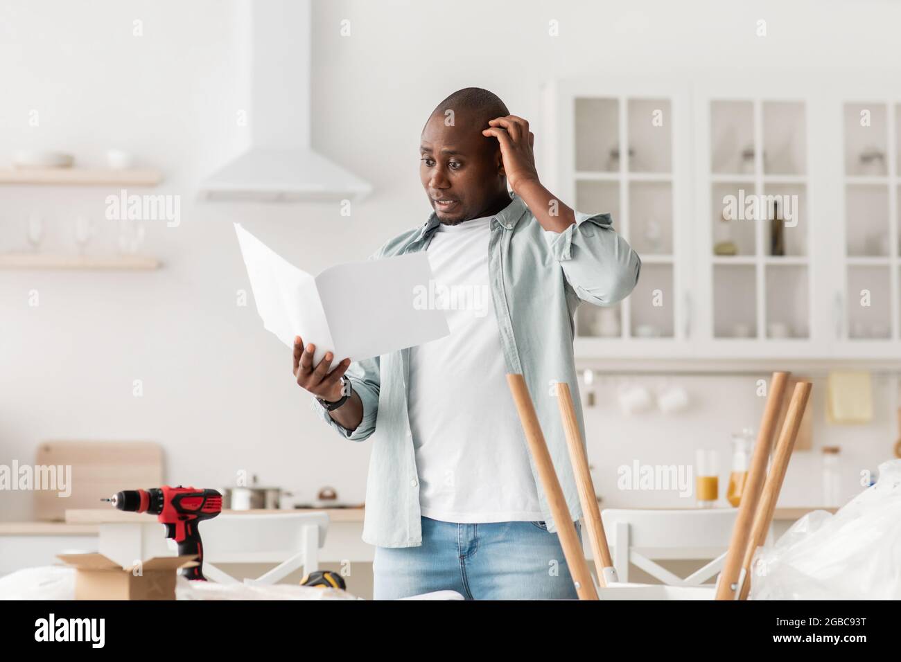 Homme noir confus ayant du mal à assembler des meubles et à lire le manuel d'auto-assemblage dans la cuisine, espace libre Banque D'Images
