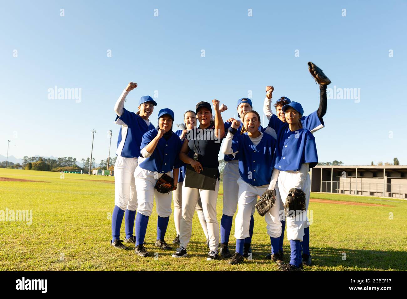 Portrait d'un groupe diversifié de joueuses de baseball et d'un entraîneur debout et applaudisant sur un terrain ensoleillé Banque D'Images