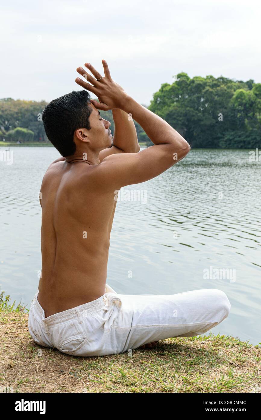 Jeune brésilien assis sur le bord d'un lac, avec ses mains opposées au-dessus de sa tête et ses pouces touchant son front. Banque D'Images