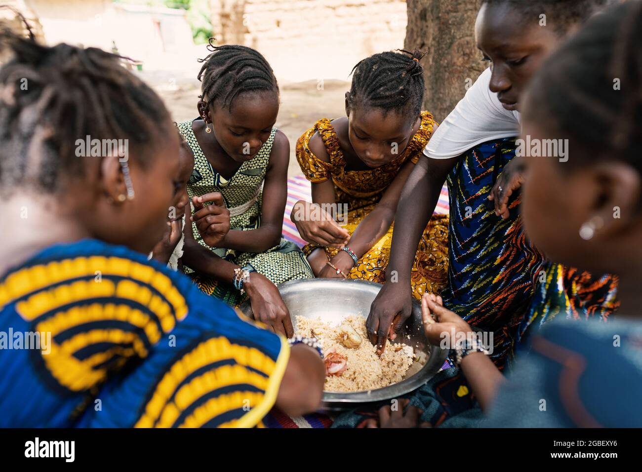 Gros plan d'une goup de filles noires habillées de coulorouille, assis autour d'un grand bol en métal, partageant un repas africain typique à base de riz et de légumes Banque D'Images